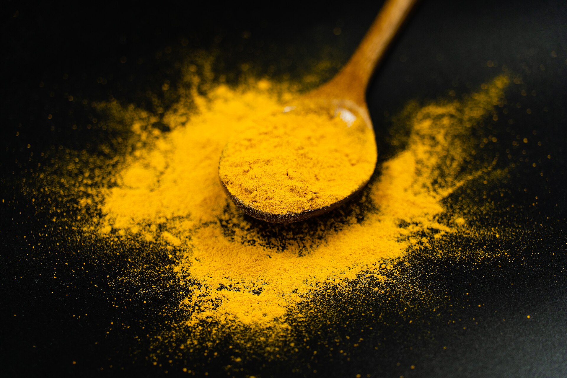 Bright yellow turmeric powder on a wooden spoon against a dark background, showing the vivid color of curcuminoids